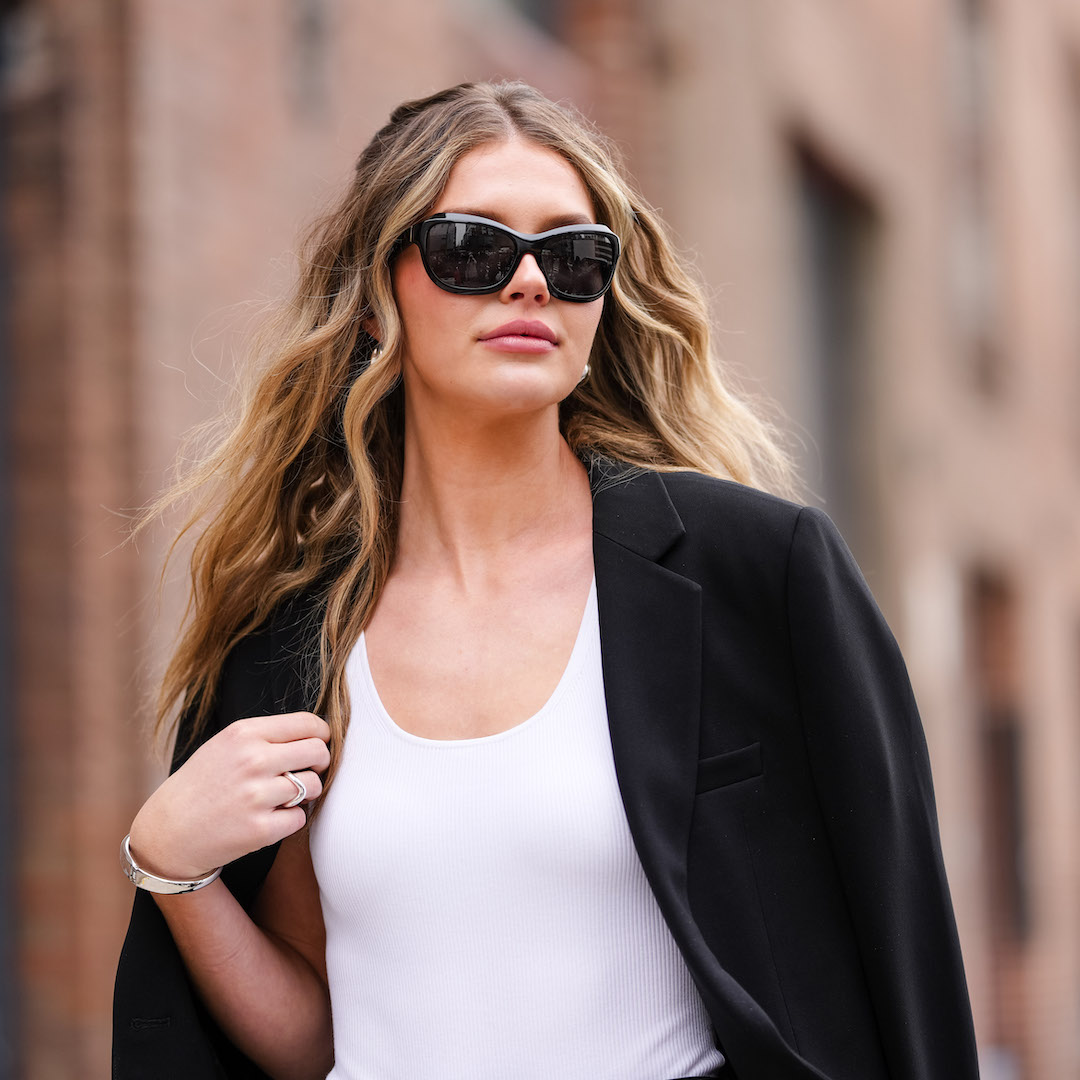 Woman with lived-in blonde hair colour wears black sunglasses, gold earrings, white tank top, black oversized blazer jacket