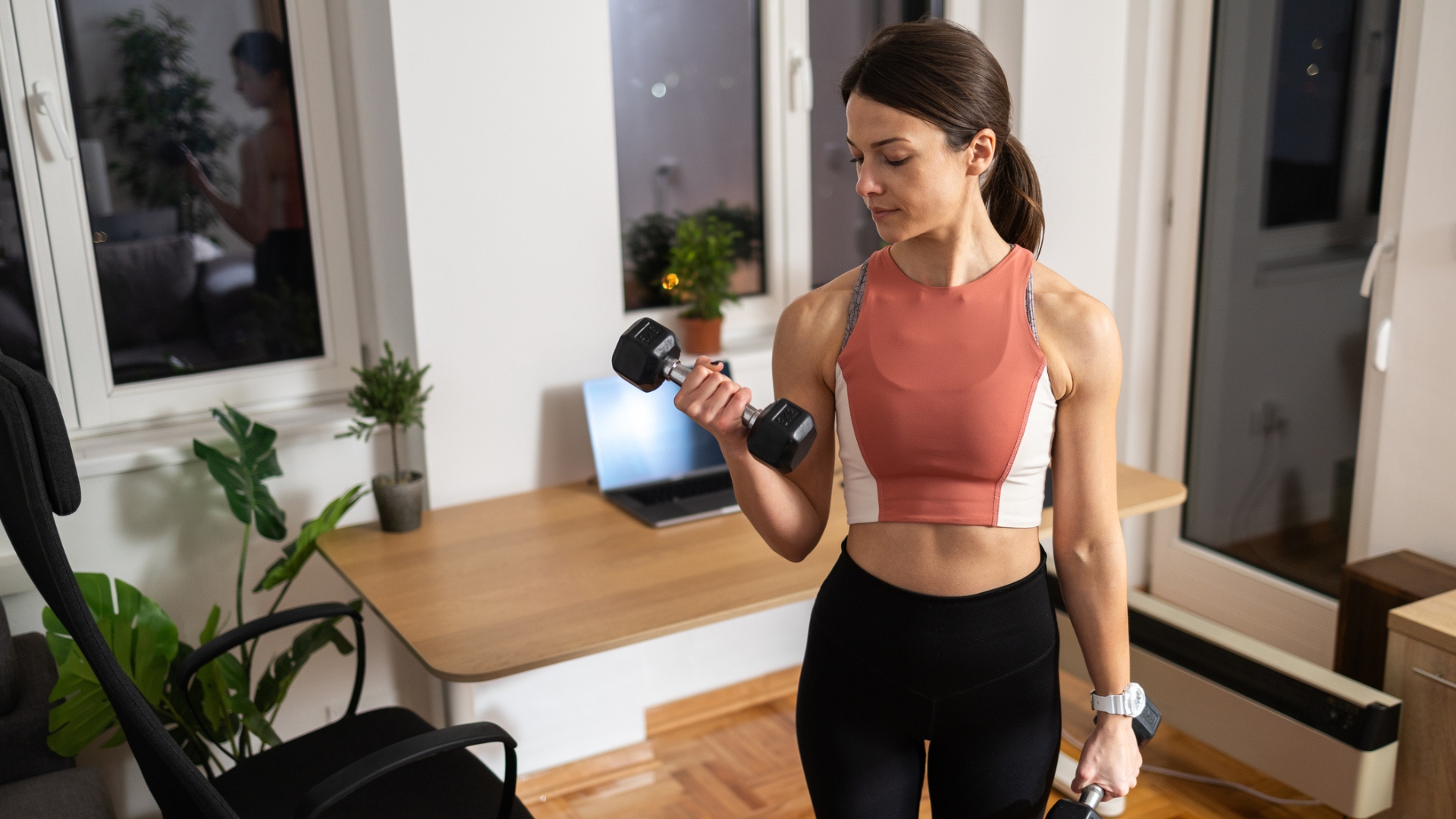 woman in a home setting holding a dumbbell in each hand, curling one up toward her shoulder and looking at it. she&#039;s wearing a coral crop top and black leggings. 
