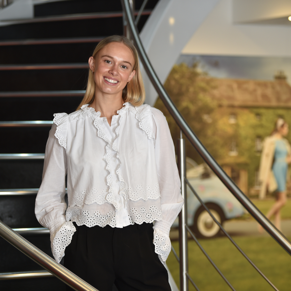 female stood on staircase with long blond hair wearing white top and black trousers