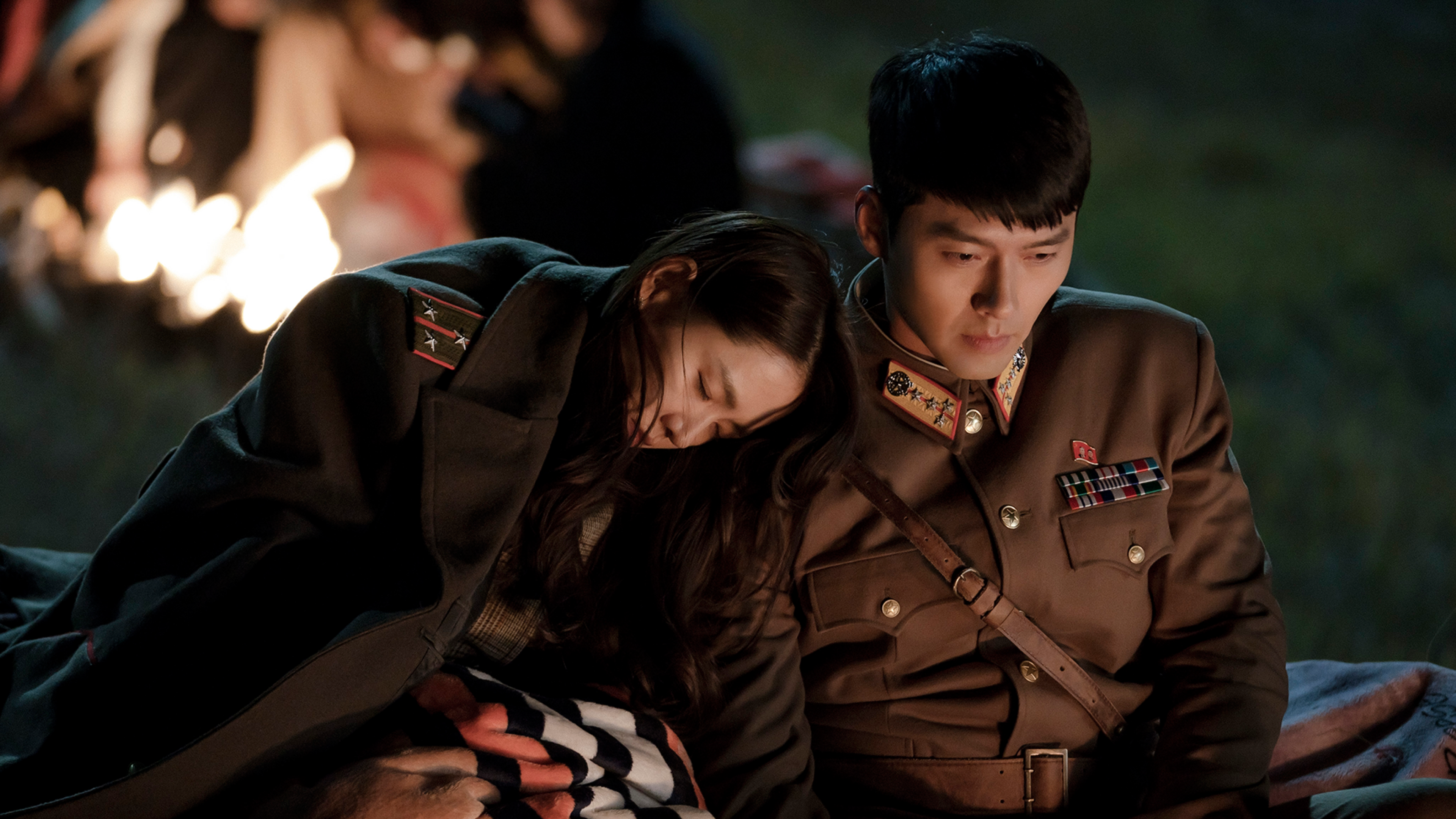 a woman wears a military jacket and rests her head on a soldier's shoulder outside of a fire in a still from crash landing on you