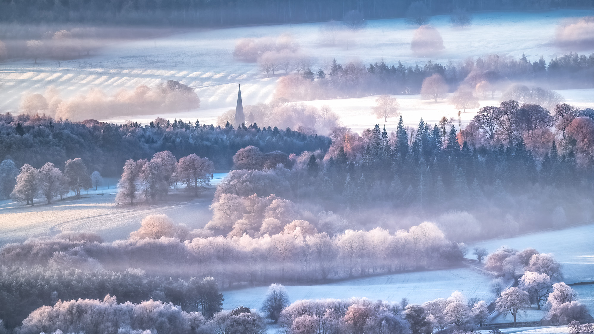 Frosty and misty landscape of the Peak District, with a church spire in the far distance surrounded by layers of open fields and forest