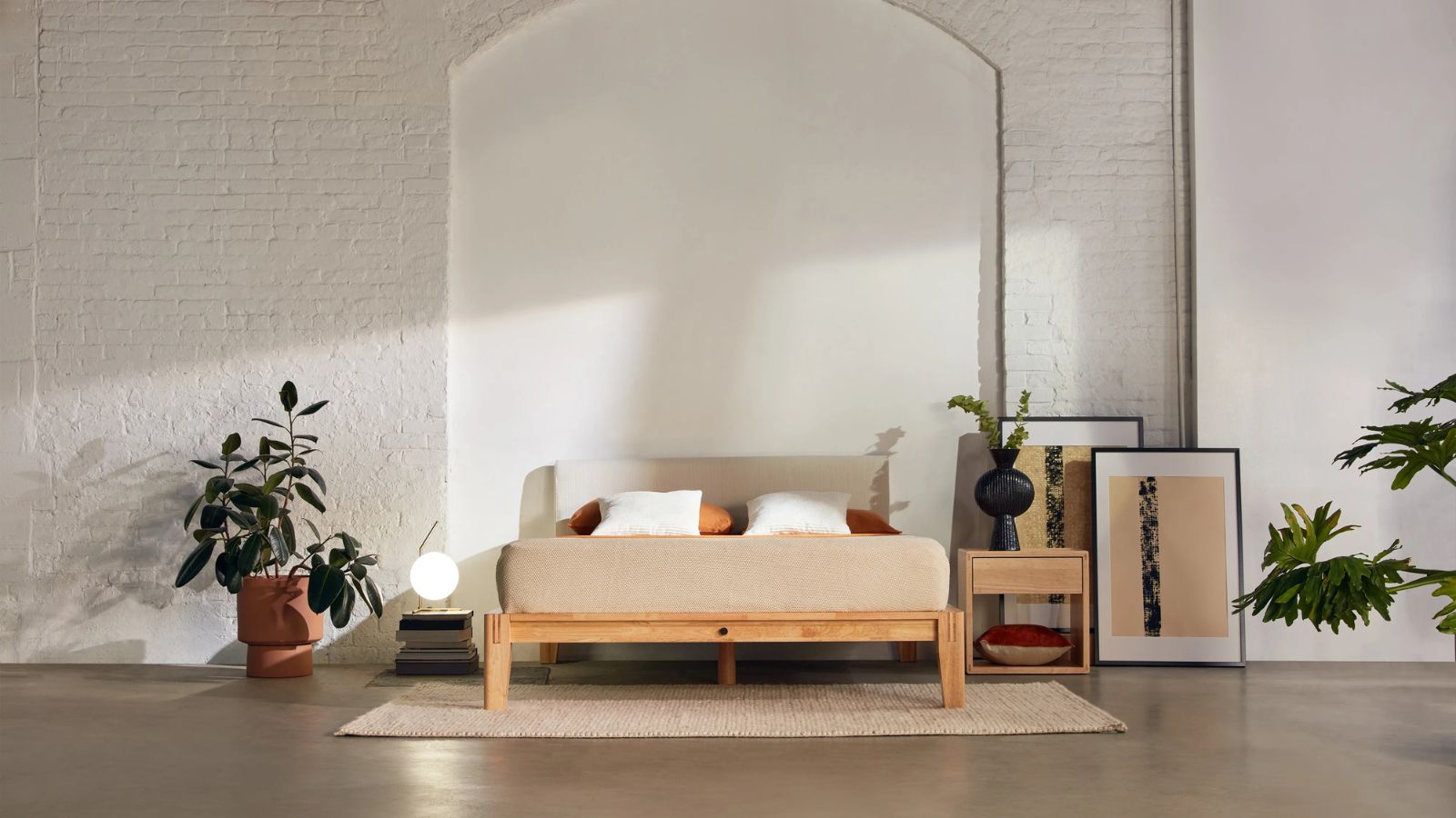 Mattress and pillows on a Thuma Bed Frame against cream walls; surrounded by potted plants, wall art and a nightstand. 
