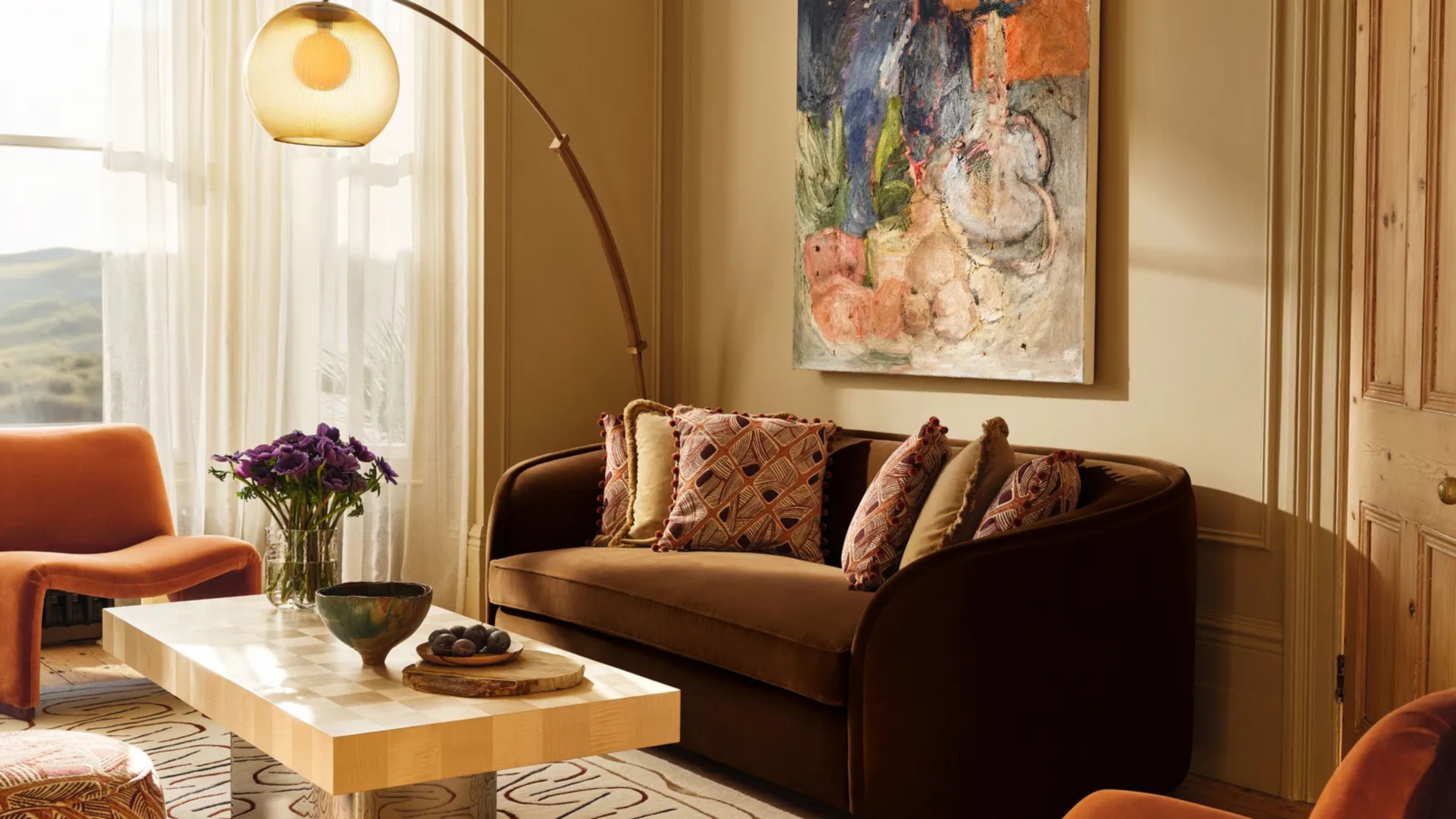Image of a beige living room with a chocolate brown sofa against the wall. In front of the sofa is a coffee table with a chrome stand and a light wooden top. There are two mid-century-style, burnt orange accent chairs and an arched floor lamp in the corner that has a bulb shade