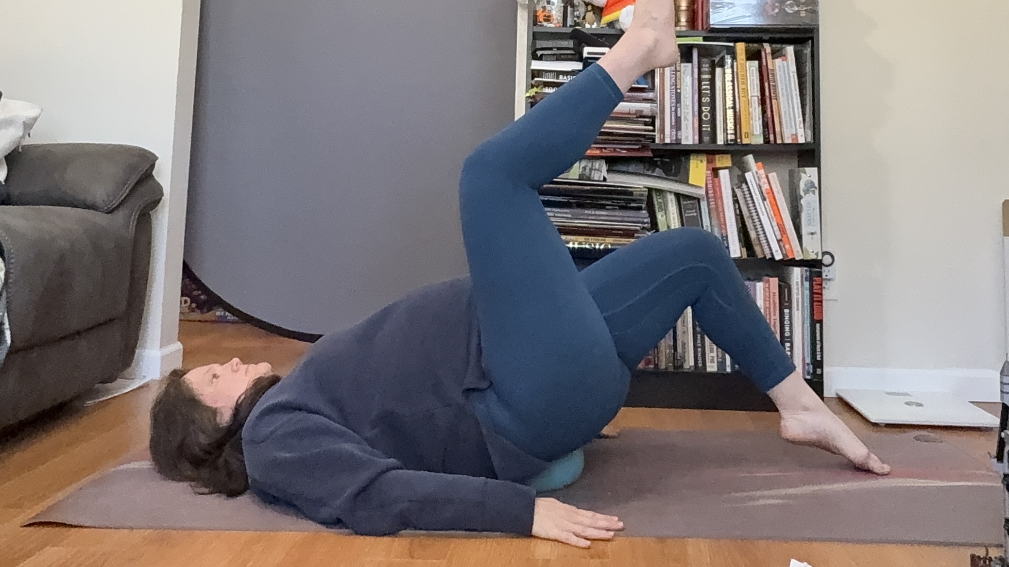 Jennifer Rizzuto performs a toe tap Pilates exercise inside on a mat. She is lying on her back with her arms by her sides and a Pilates ball placed underneath her hips. Her knees are bent, with one leg in the air and the other tapping on the floor.
