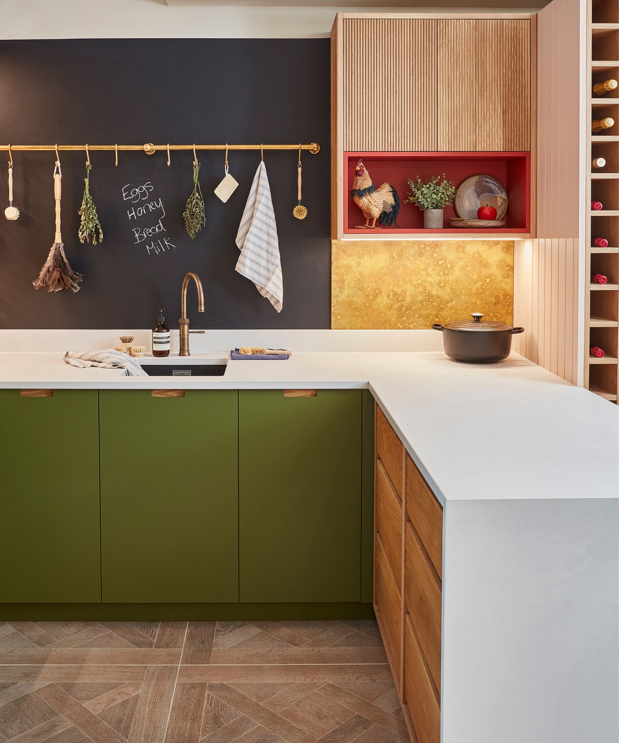 A modern kitchen with deep green lower cabinets, white countertops, and a black chalkboard wall with a brass rail holding kitchen essentials. A rooster figurine sits in a red shelf nook, and a wine rack is built into the cabinetry.