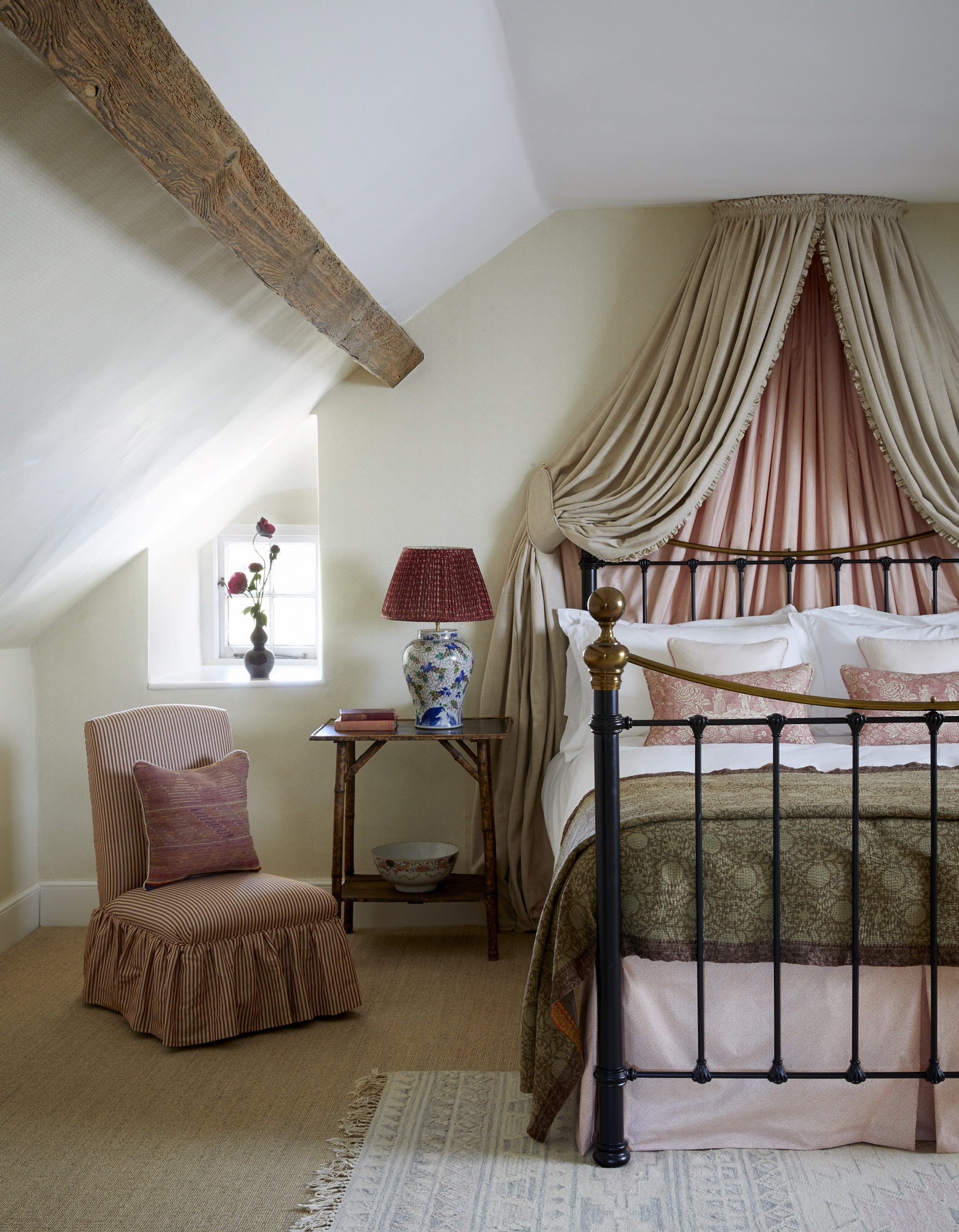 attic bedroom with canopy, iron bed, coir carpet, upholstered chair, side table, rug, beam, pinks