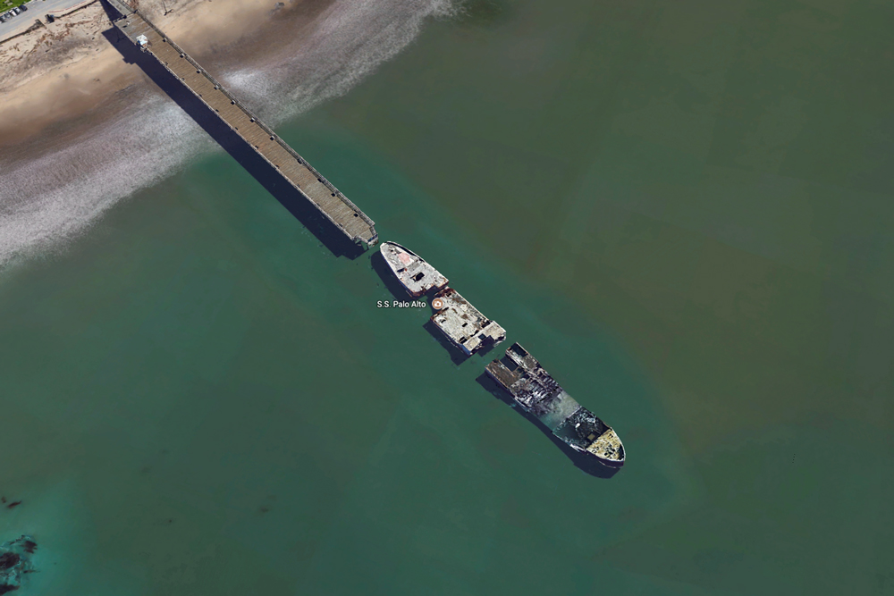 We see a bird's-eye view of a long broke military ship floating at the end of a dock on a beach.