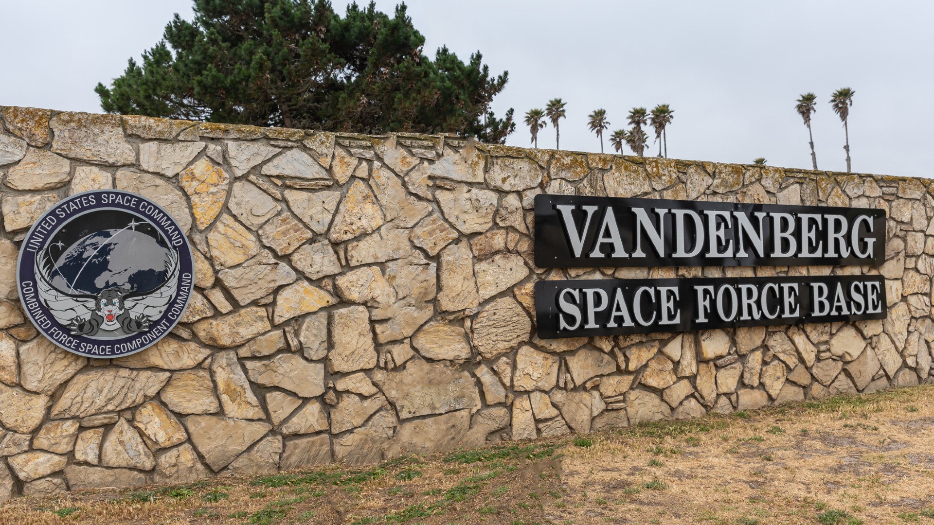 a stone sign reading "vandenberg space force base" with tall palm trees visible behind it
