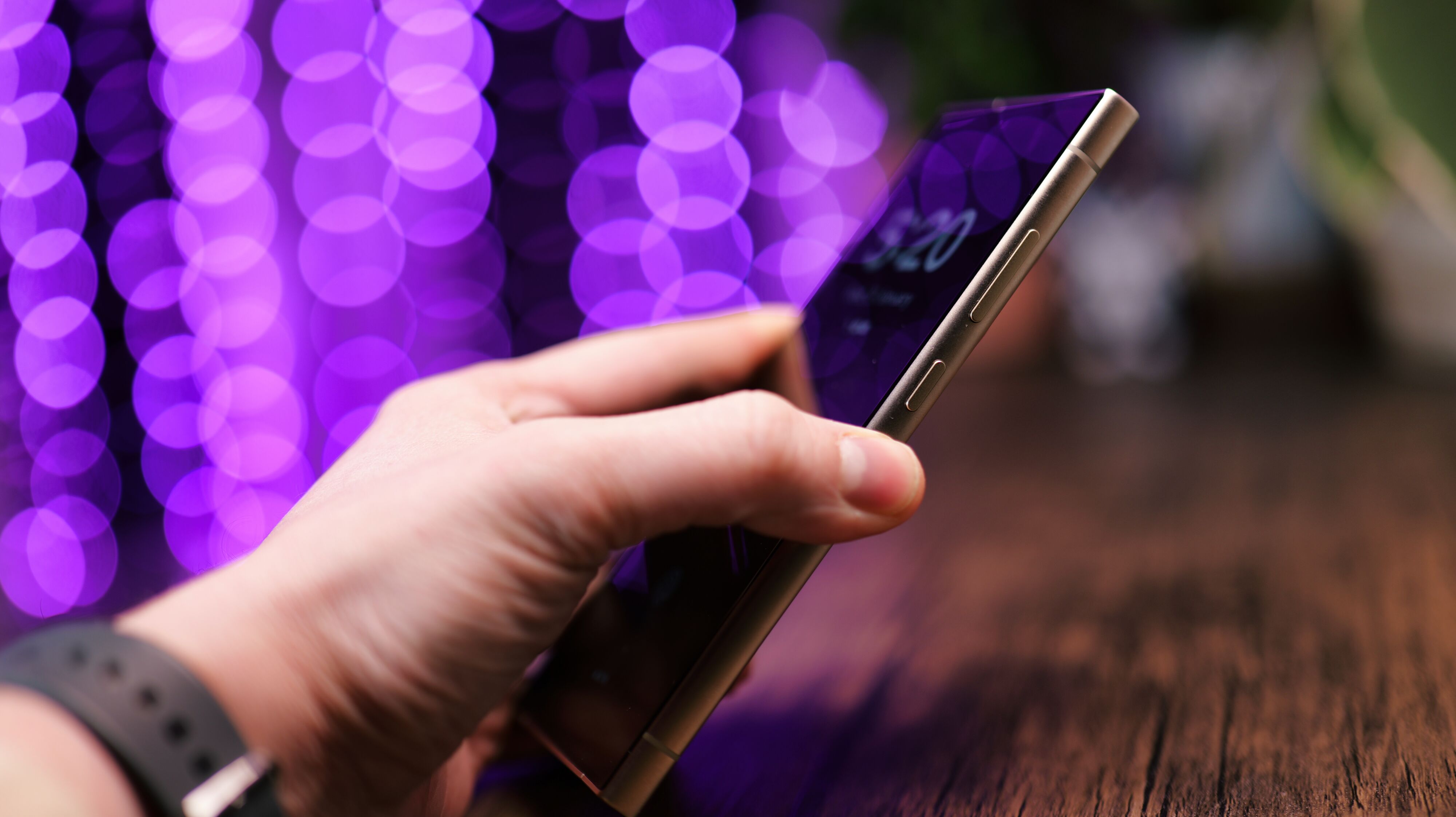 Side of a Samsung Galaxy S24 Ultra phone held in a hand with purple lights blurred in the background
