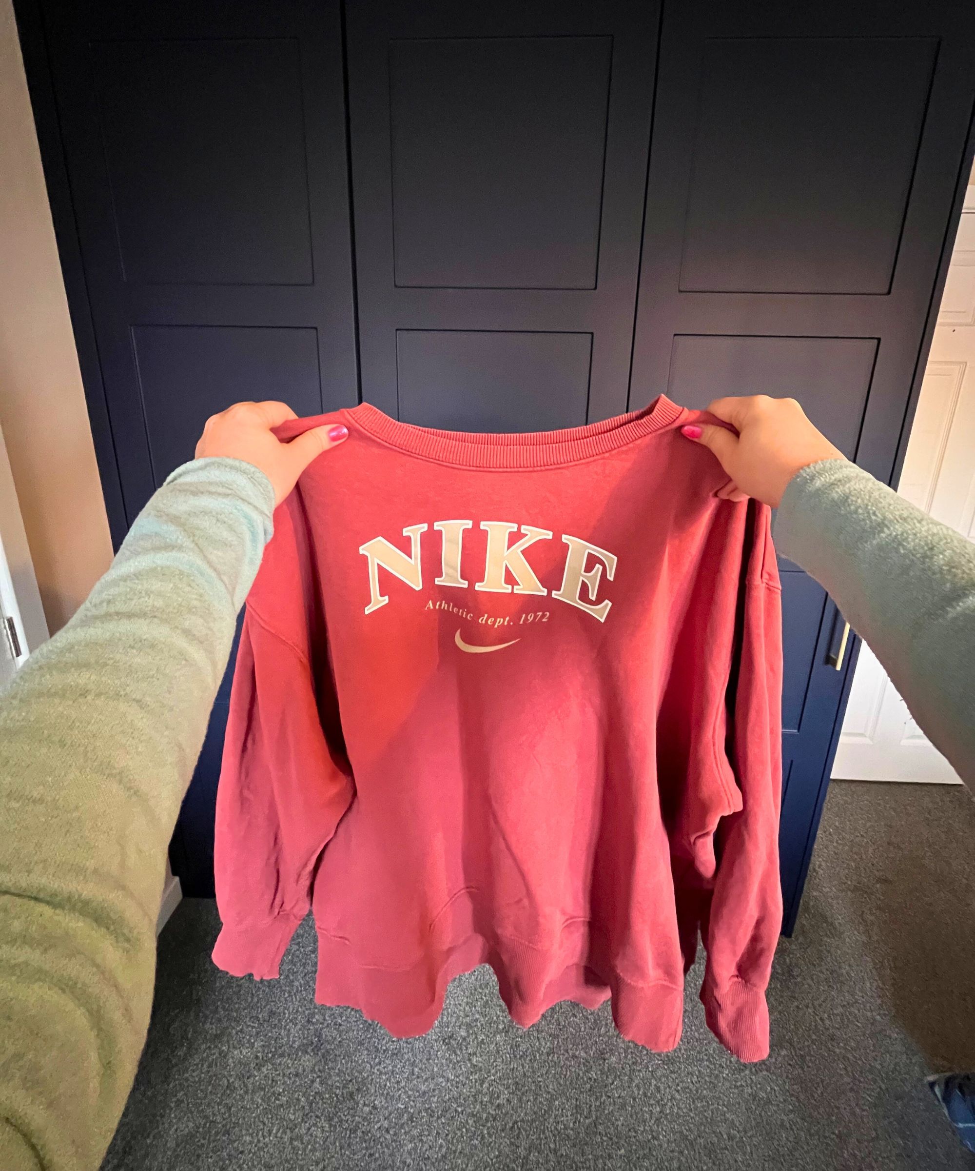 The image shows a point of view shot of a peachy-orange Nike long sleeve sweater being held in front of the camera with thumbs either side of the neck