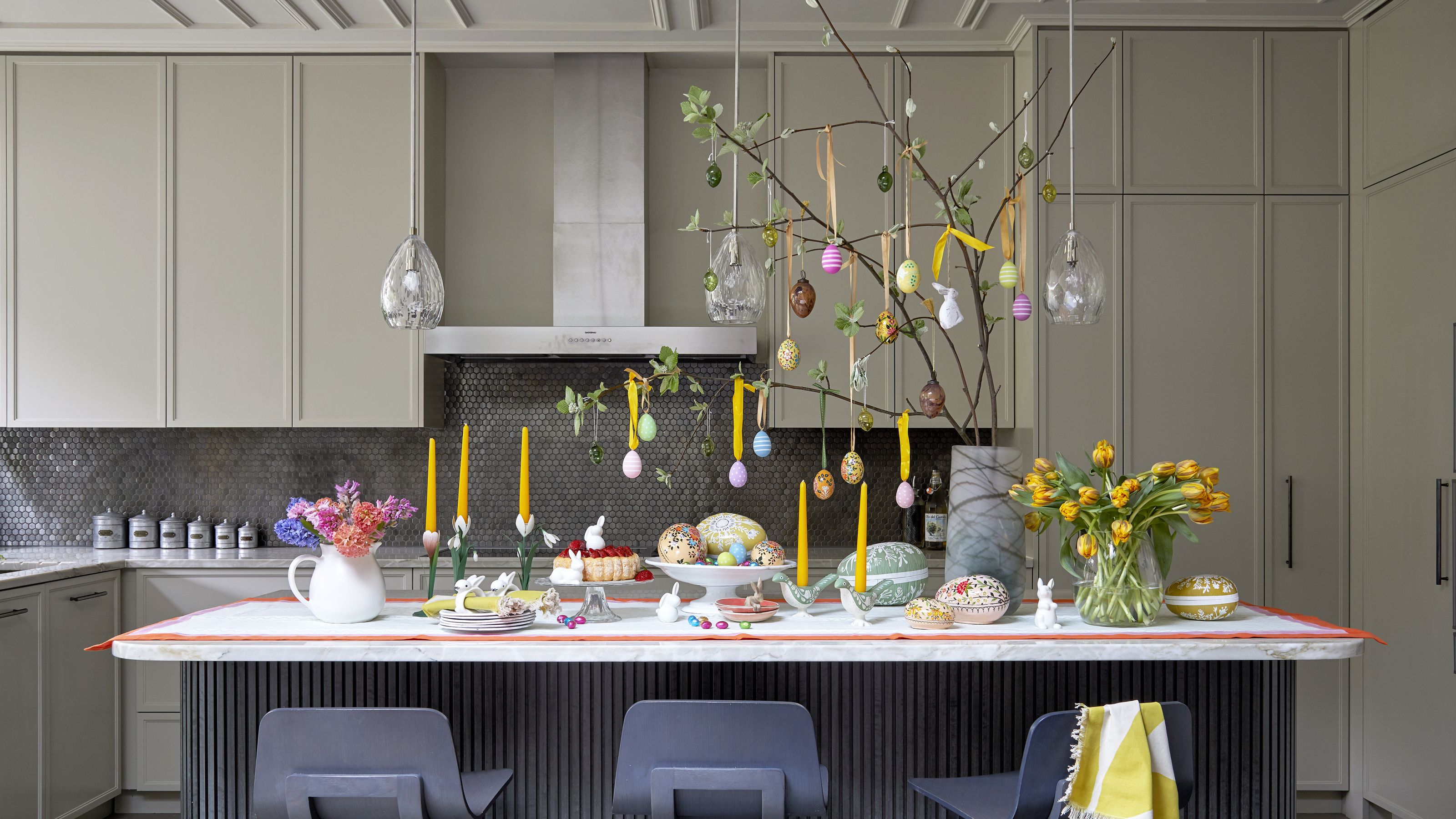 A kitchen island dining table decorated for Easter with taper candles, tulips, and egg decorations hanging from twigs of willow