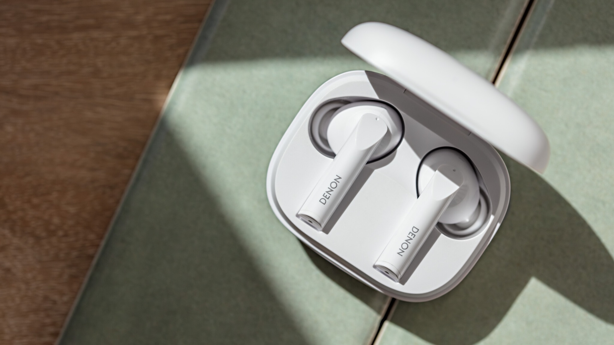 Denon AH-C840NCW true wireless earbuds in white, in their charging case on a green fabric and wooden surface