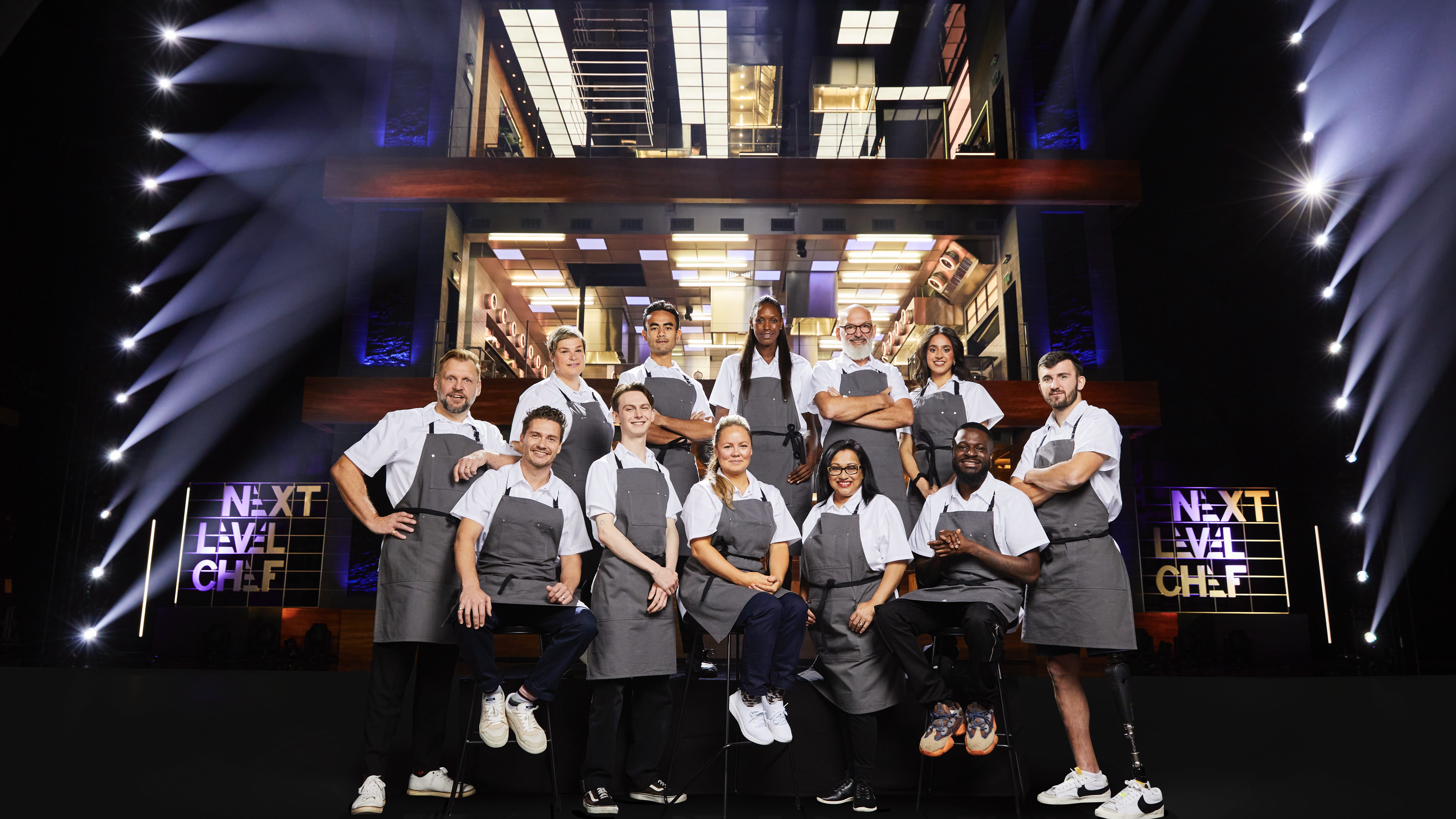 The contestants wearing white tops and grey aprons stand in a group in front of the three-storey kitchen in Next Level Chef.