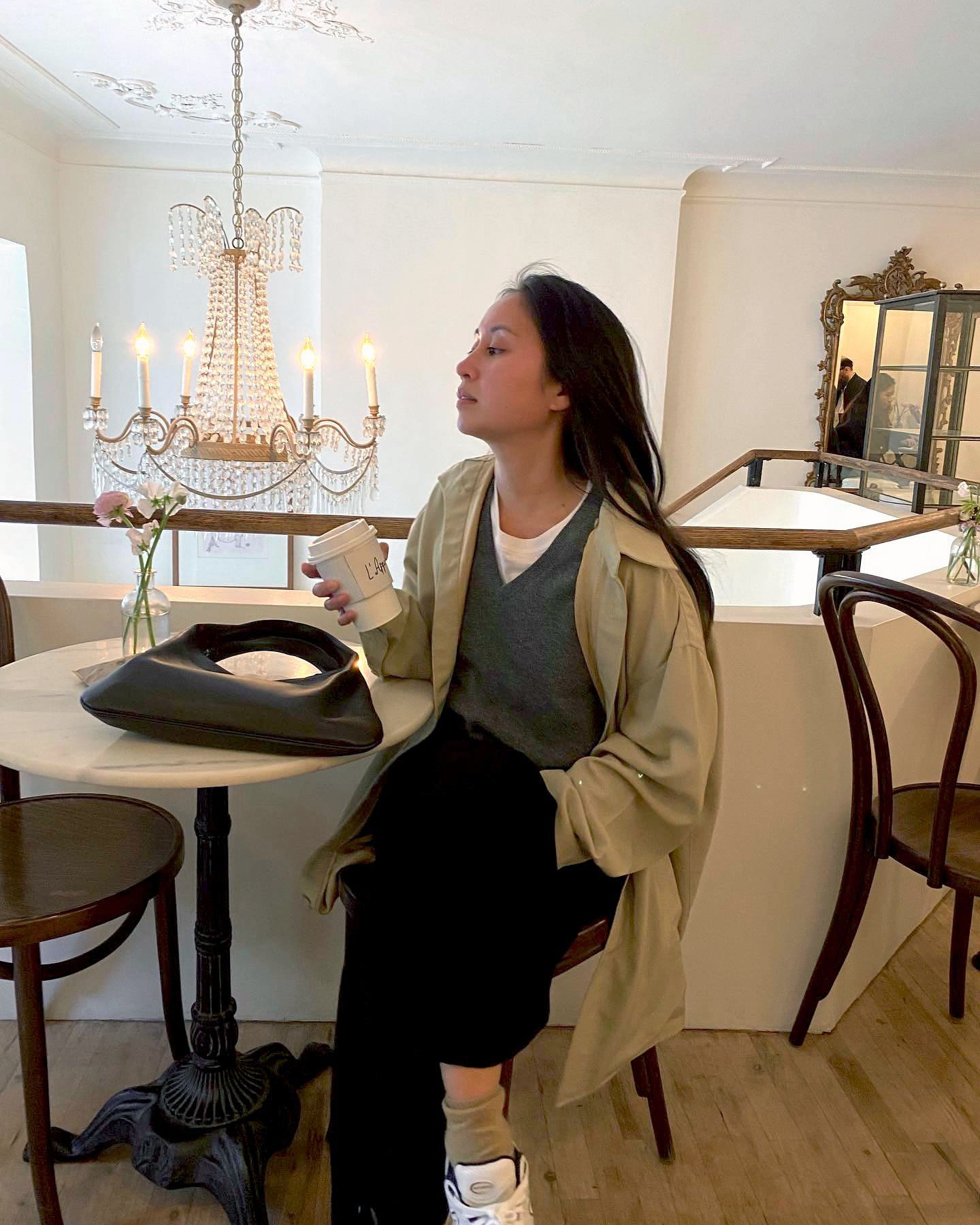 Jordan Risa Santos wearing a tan trench coat over a gray v-neck sweater, a white tee, and black trousers.