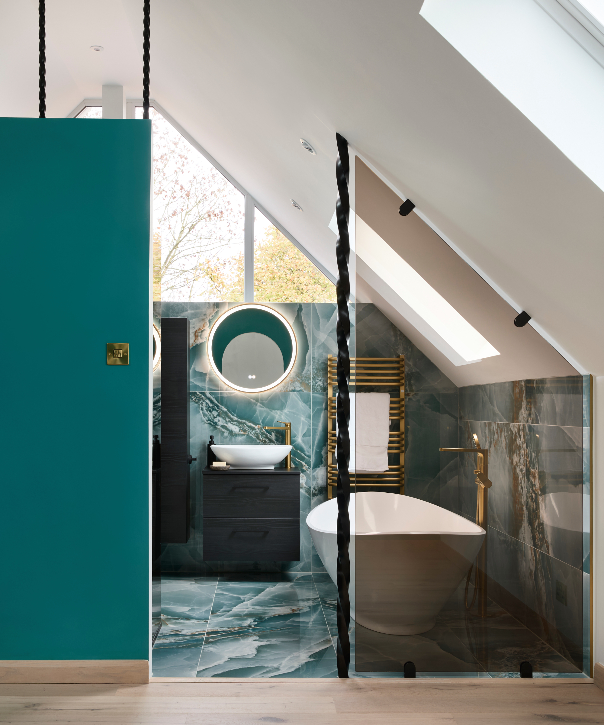 bathroom with blue marbled floor and wall tiles, roof light, white freestanding bath, blue wall outside room and twisted ornate wooden spindles on 3/4 height wall and to side of glass screen on one side of entrance