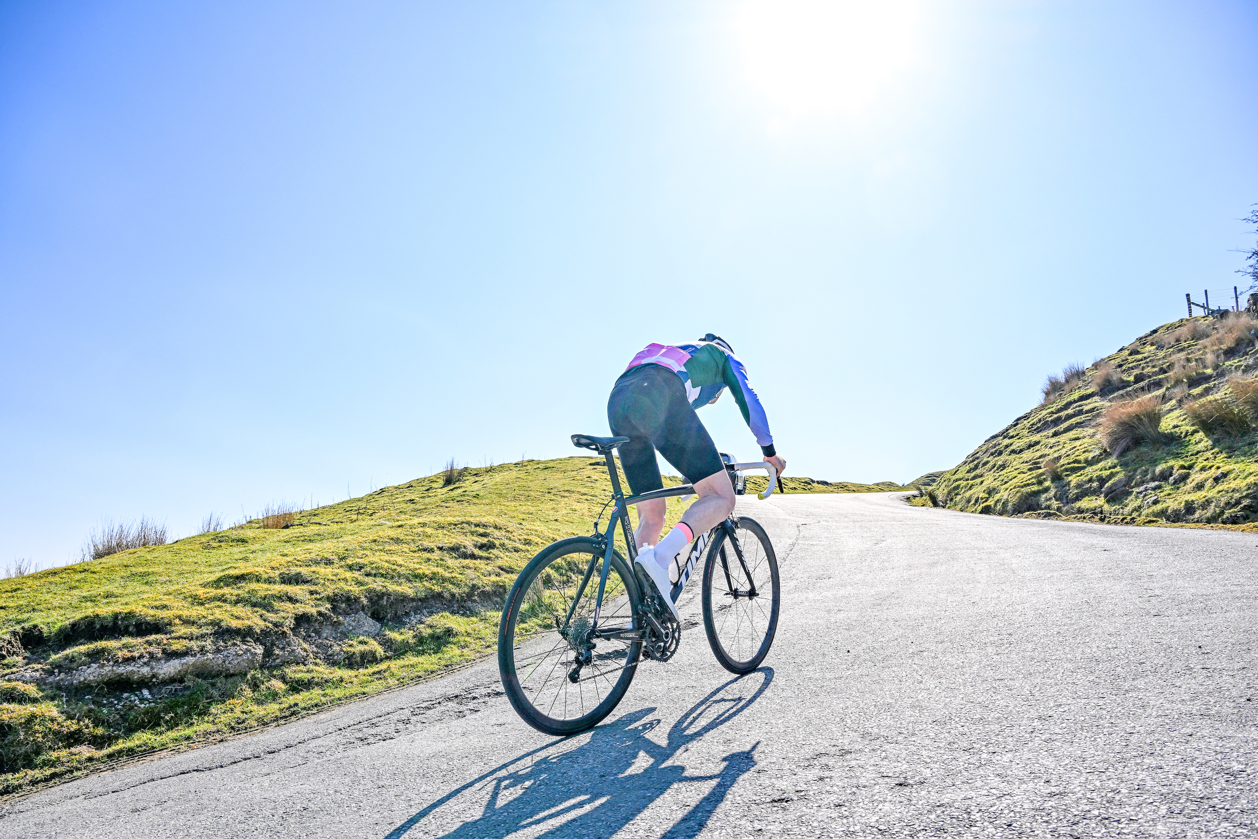 A rider climbing a hill, blue kit, blue sky, glaring sunlight, with the sun directly overhead and almost in shot