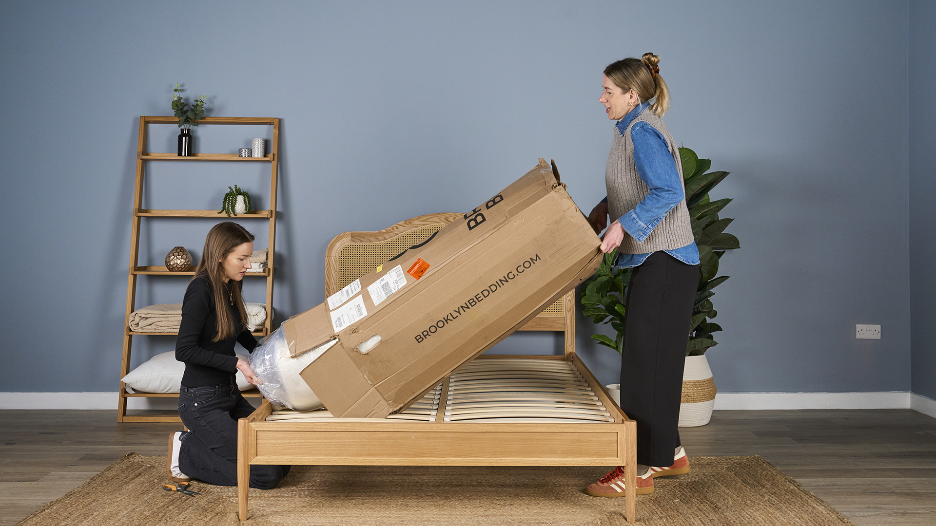 Two girls unboxing the Brooklyn Bedding CopperFlex mattress in sleep studio with rattan bedframe, shelf and plants