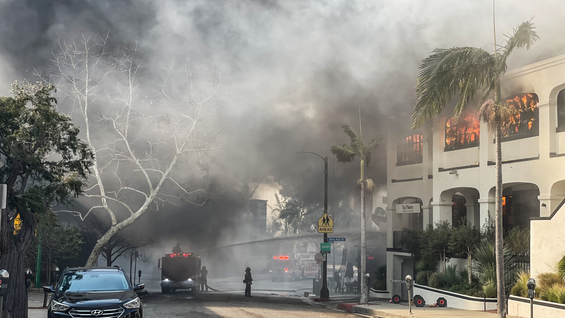 A picture of a street whereby houses are burning as a result of wildfire smoke. Grey and black smoke can be seen covering half of the image.