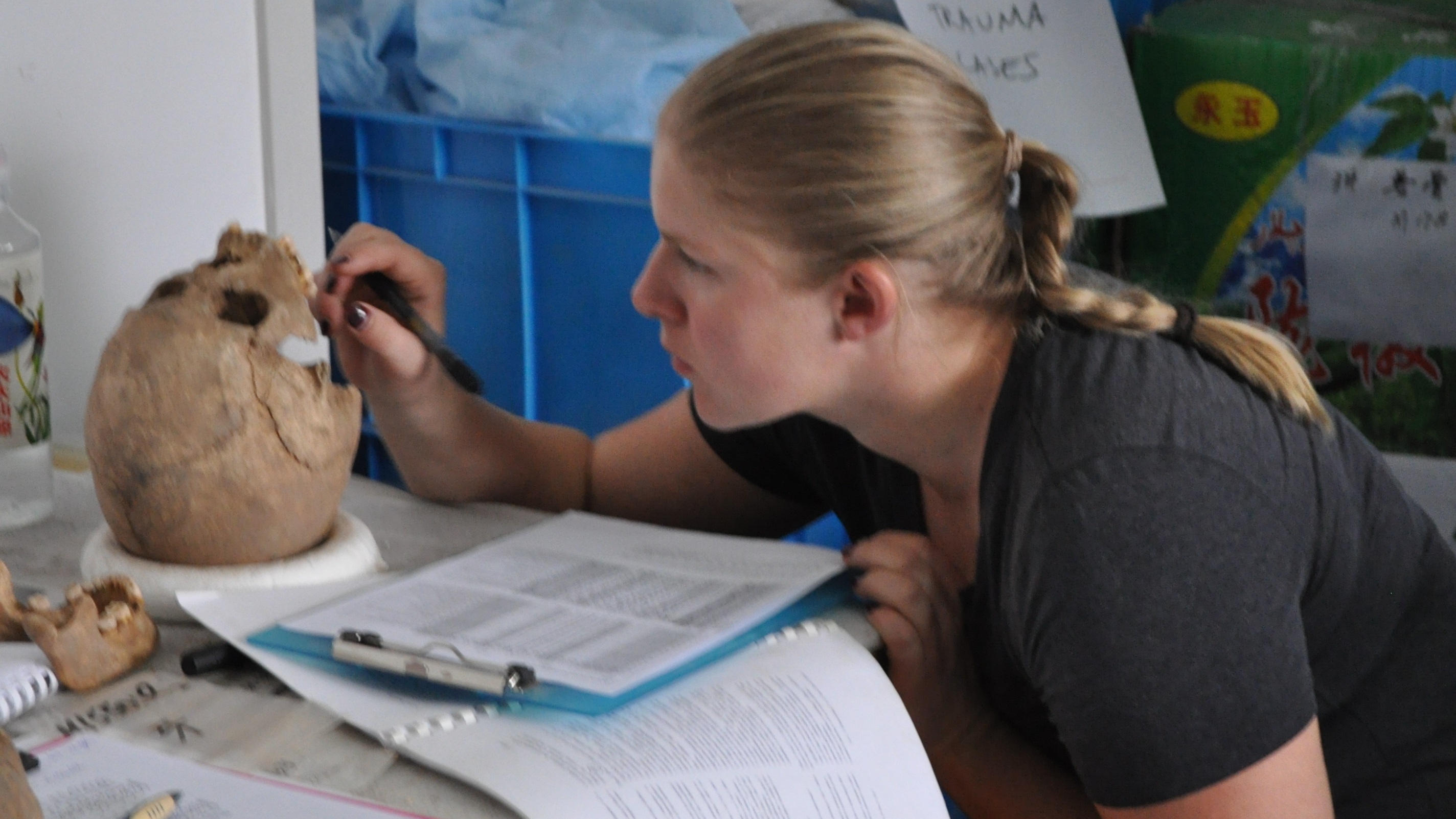 A white woman with blonde hair in a ponytail looks at a human skull on a table
