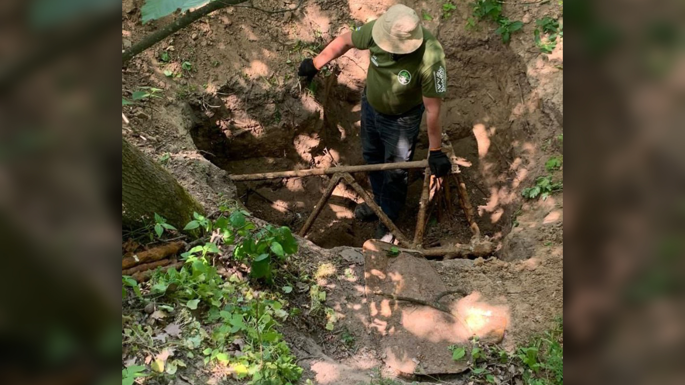 We see a man in a hat in a dirt hole and holding onto part of a rusty, decayed plane.