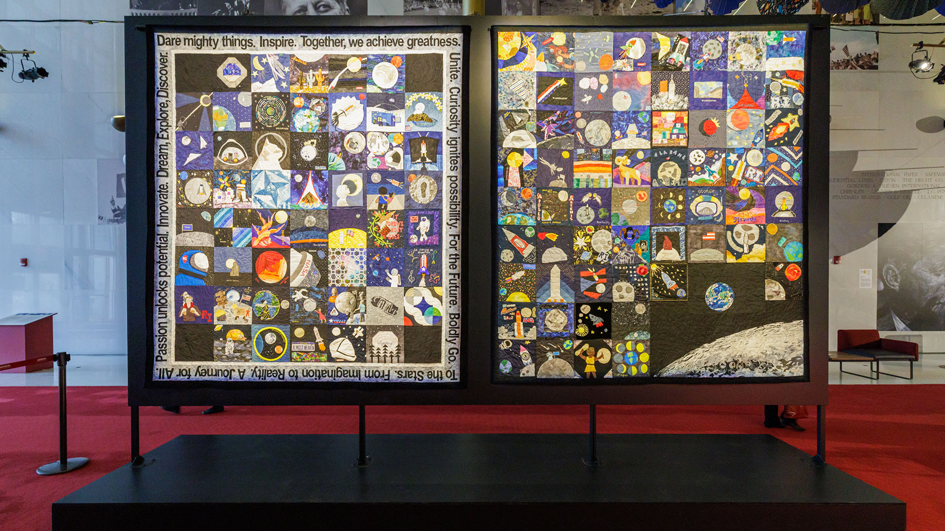 two colorful patchwork quilts are framed and on display in a red carpeted hallway