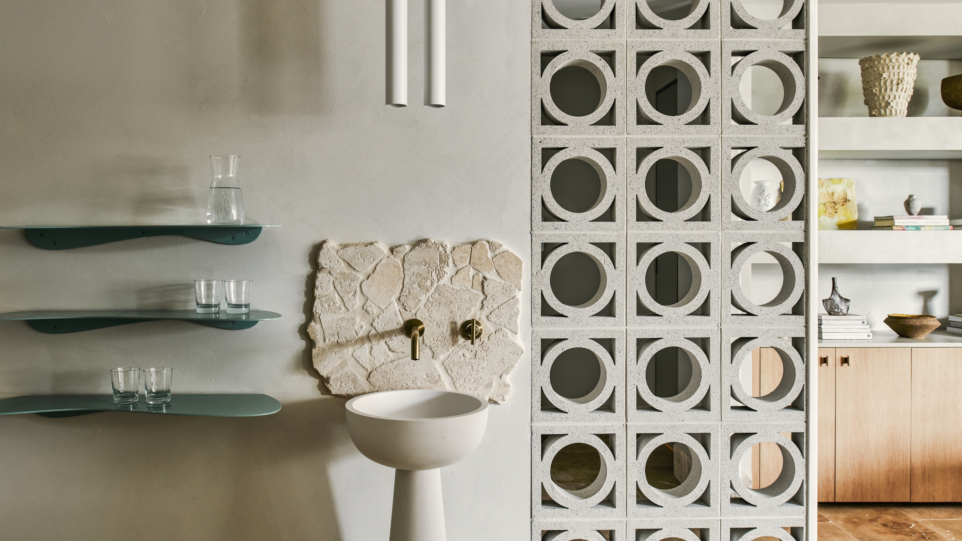 Image of a gray stone wall with a stand alone sink. Beside the sink are three blue, mini floating shelves with glasses on them