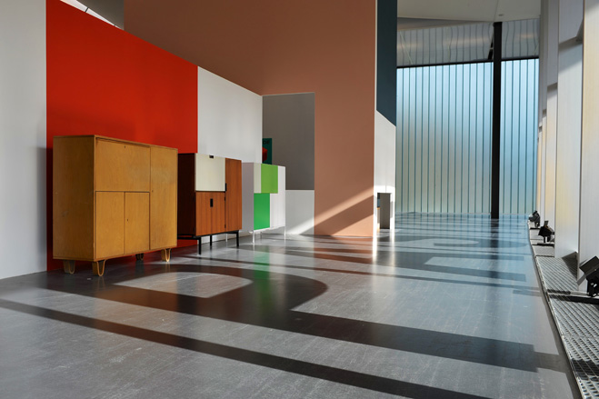 An exhibition space with three multi-compartment cabinets, one in wood/brown, one in red and white and one in green and white. 