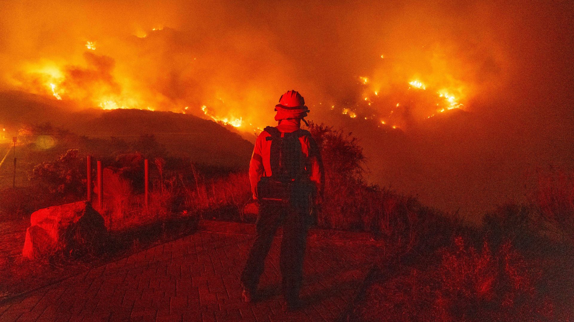 a firefighter wearing gear stands on a hill looking out at a large wildfire