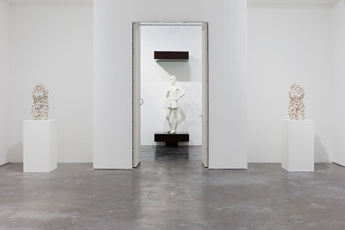 Two small statues are set on both sides of a door through which we see a white statue of a woman.