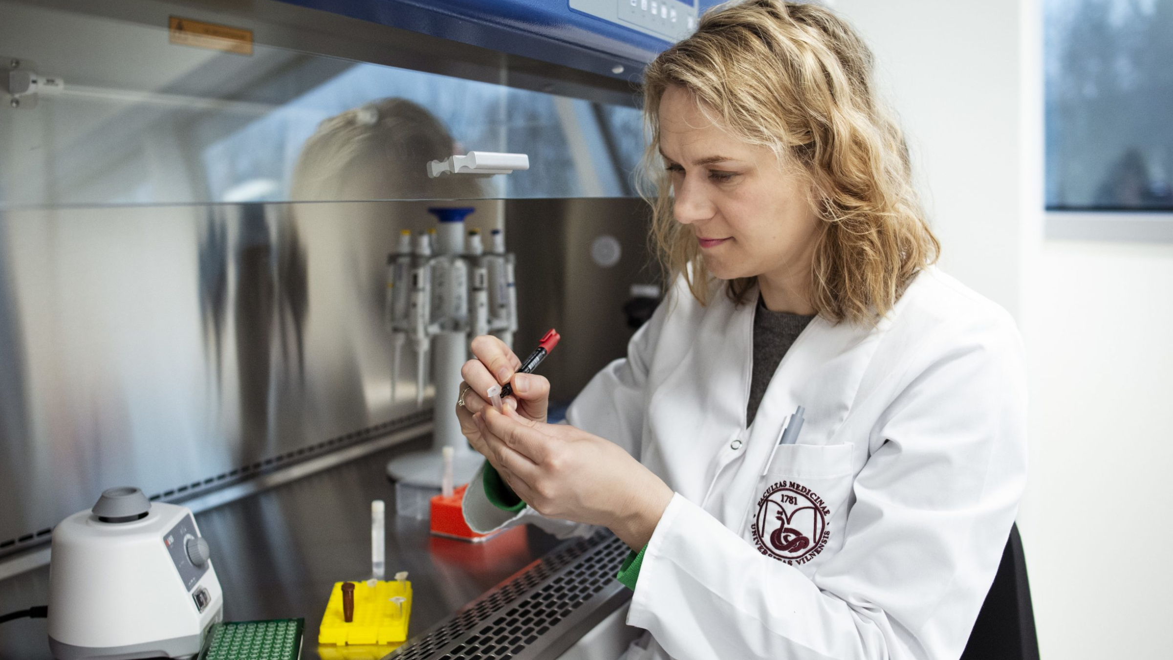 A picture of Ingrida Domarkienė sat at a lab bench using a marker to write on a test tube. She is wearing a white lab coat. 