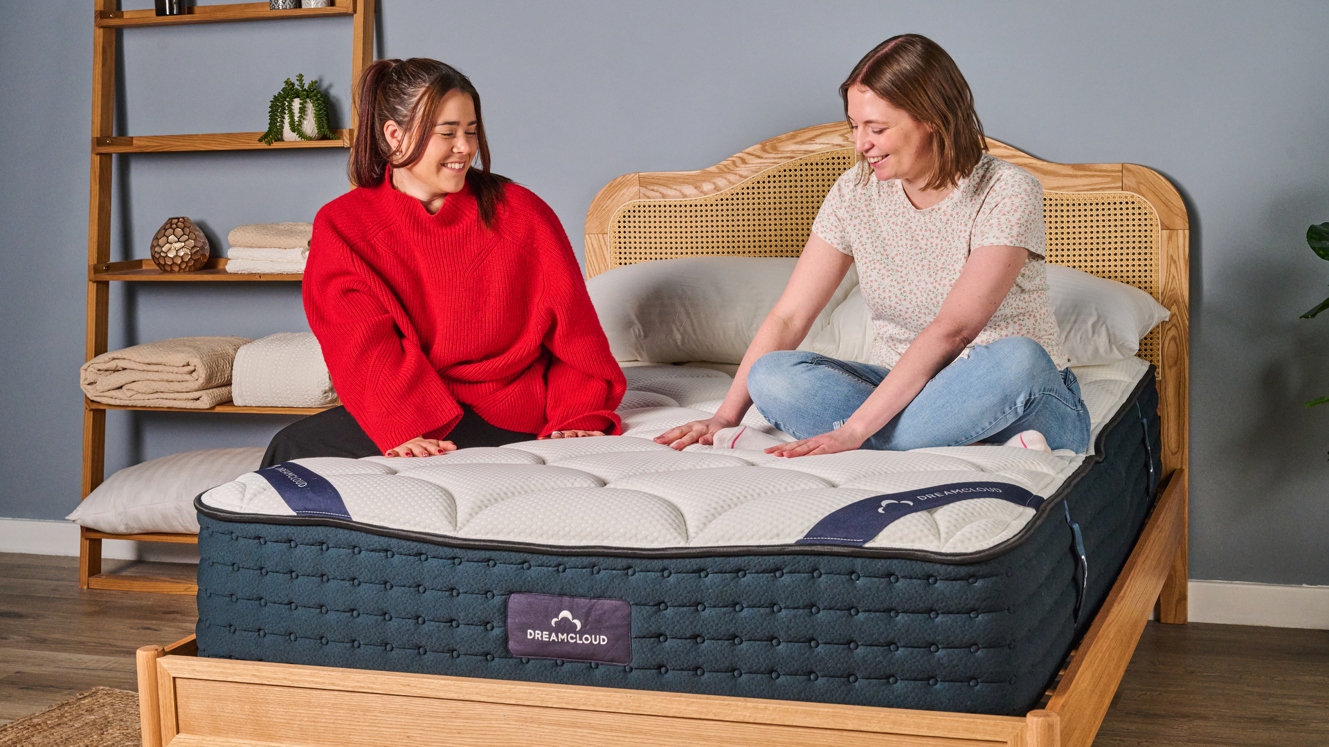The image shows two women sitting on top of the DreamCloud Hybrid Mattress on a wooden bed frame in a blue bedroom