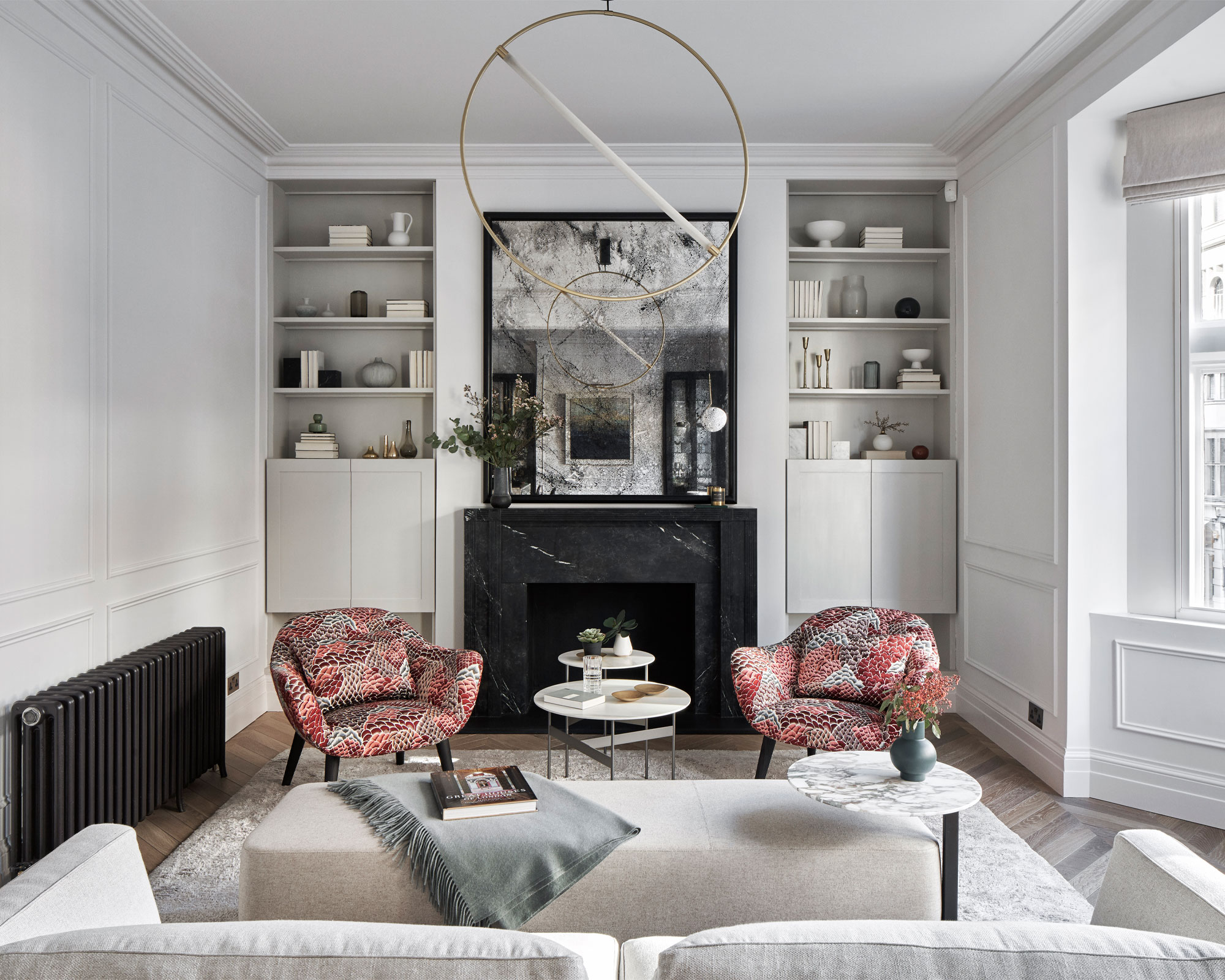 Living room with pale grey walls, bay window, built in storage either side of the chimney breast and fireplace. Grey rug and sofa and orange patterned armchairs.