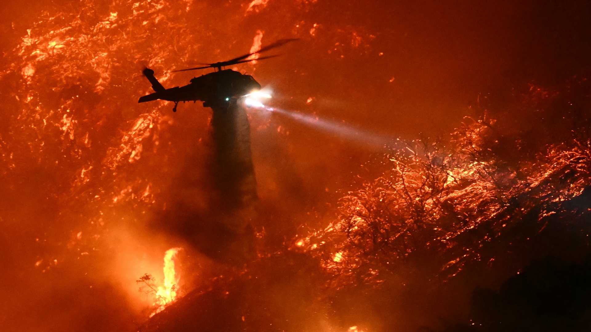A helicopter drops water over a raging wildfire.