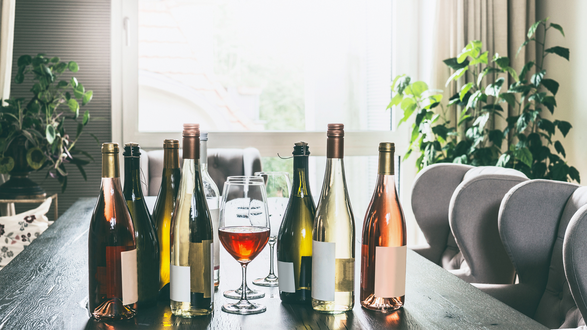 Variety of wine bottles with mock up label and glasses on table in modern living room at window.