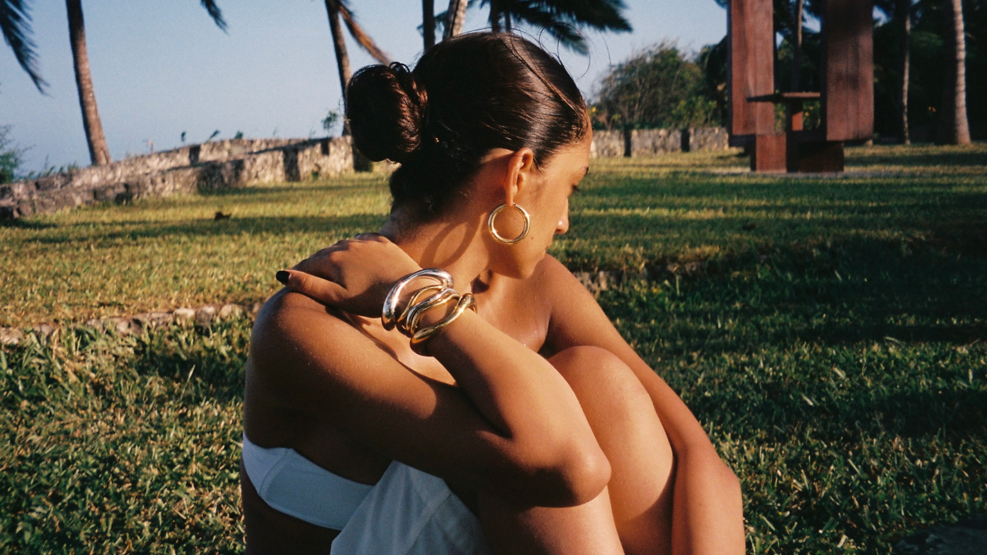 Content creator Emna Sellimi sitting in the grass wearing a white bralett, skirt, and a stack of silver bangle bracelets